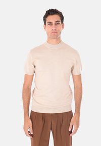 Uomo in piedi, indossa una maglietta a maniche corte beige in maglia e pantaloni marroni con pieghe a vita alta, guarda davanti a sé con un'espressione neutra.