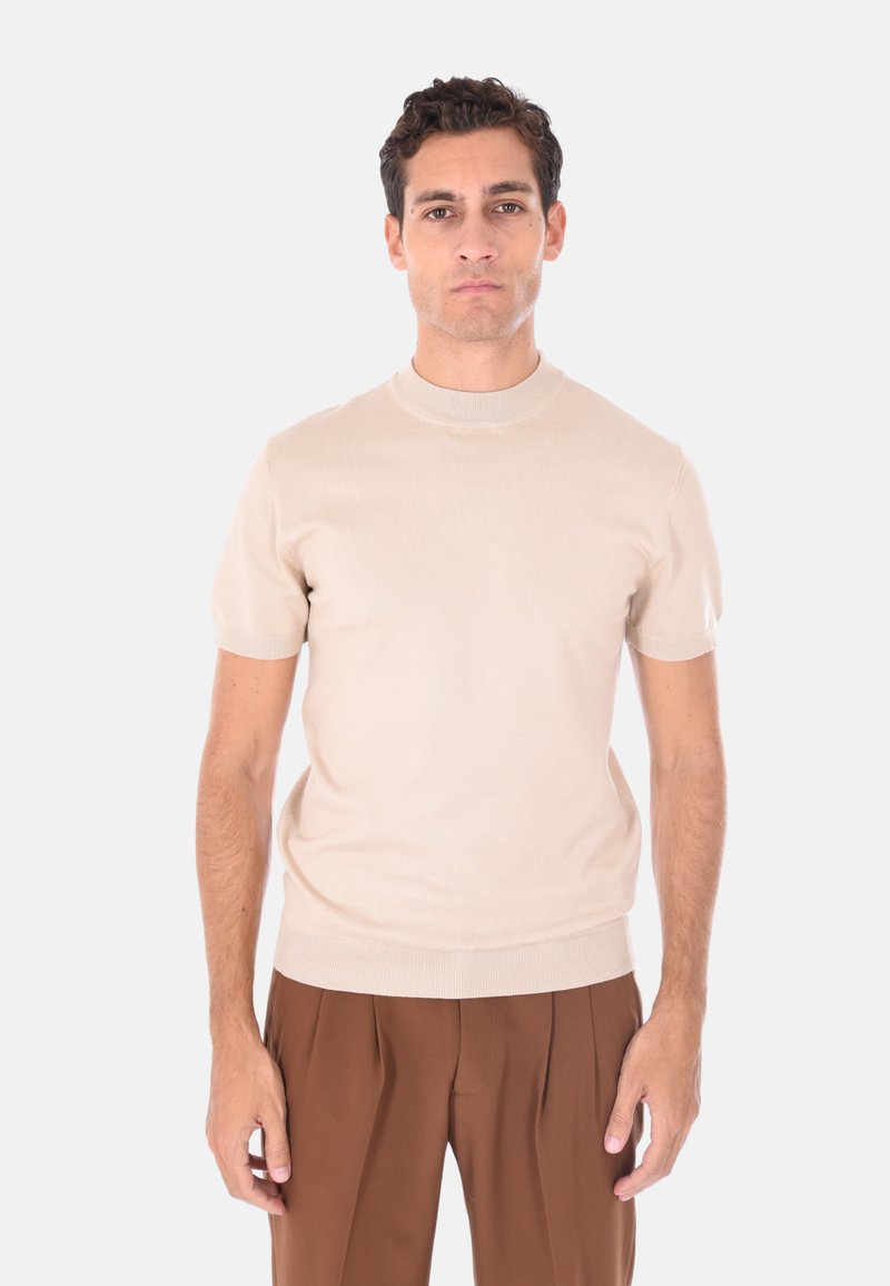 Uomo in piedi, indossa una maglietta a maniche corte beige in maglia e pantaloni marroni con pieghe a vita alta, guarda davanti a sé con un'espressione neutra.