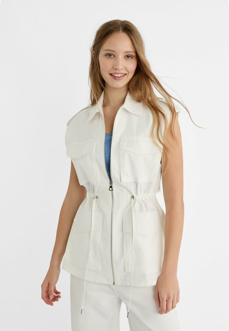 Femme souriante avec de longs cheveux portant un gilet sans manches blanc à fermeture éclair avec de grandes poches et un pantalon blanc sur un fond uni.