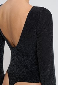 Czarny bodysuit z długim rękawem i głębokim dekoltem z tyłu, wykonany z fakturowanego, błyszczącego materiału o dopasowanym kroju. Podkreślony subtelnym połyskiem.