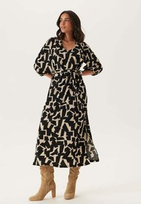 Abito lungo nero e beige con motivi geometrici, dotato di vita aderente, maniche a palloncino e uno spacco laterale. Abbinato a stivali al ginocchio beige.