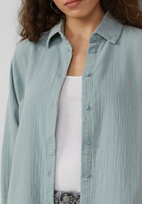 Lyseblå tekstureret skjorte med knapper og krave, seks knapper foran; båret over en hvid top.