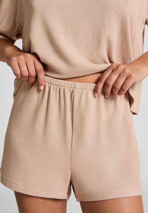 Persoon die de elastische tailleband van beige gebreide shorts aanpast, gekleed in een bijpassend beige gebreid topje, met zichtbare buik en handen.