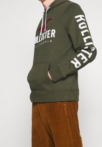 Person som bär en olivgrön Hollister-hoodie med vitt logotyp och text på ärmen, kombinerat med bruna manchesterbyxor, st ående på sidan.