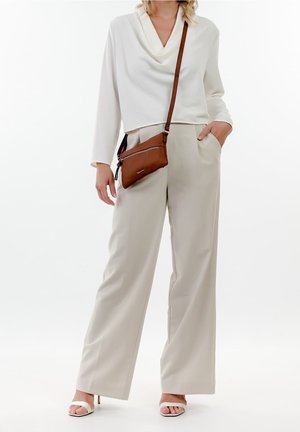 Kobieta ubrana w biały długi rękaw, beżowe spodnie o szerokich nogawkach, białe sandały na obcasie oraz brązową torebkę na ramię.