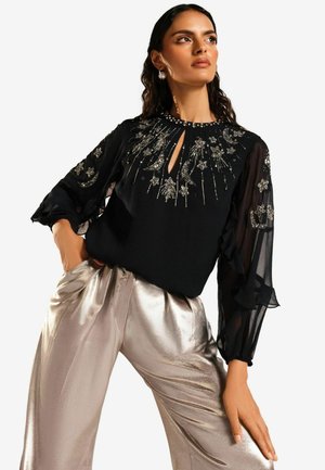 Svart blus med genomskinliga långa ärmar och blommig broderi; har en nyckelhålsutskärning. Kombinerad med glänsande silverfärgade vida byxor.