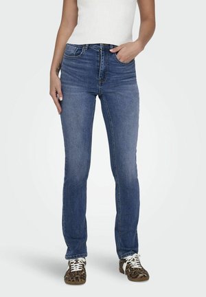 Kvinde iført blå straight-leg jeans, hvid ærmeløs top og leopardprintede sneakers, stående med den ene hånd i lommen.