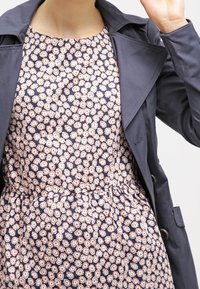 Kobieta w granatowej sukience w kwiaty i ciemnoszarym płaszczu, podnosząca prawą rękę w pobliżu głowy.