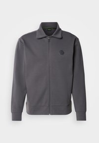 Giacca grigia con zip realizzata in tessuto liscio, caratterizzata da un alto colletto, polsini a coste e un logo esagonale sul lato sinistro del petto.