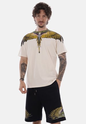 Uomo con capelli ricci e tatuaggi che indossa una camicia crema e pantaloncini neri, entrambi con motivi di ali di uccello gialle e verdi.