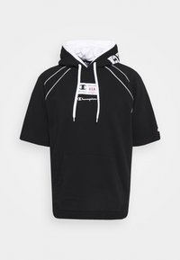Felpa nera con cappuccio e maniche corte, cuciture bianche, tasca frontale e accenti del logo. Il tessuto appare morbido e texturizzato.