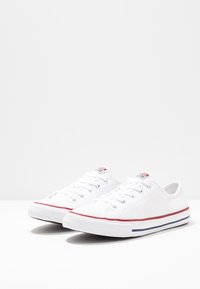 Paire de baskets basses en toile blanche avec des rayures rouges et bleues sur des semelles en caoutchouc, posées sur une surface blanche.