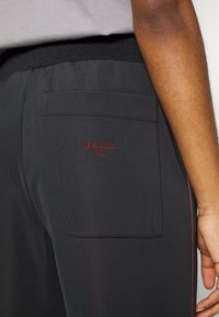 Black athletic pants with ribbed waistband, featuring a back pocket. "BALLY 1851" in red embroidery on the pocket. Textured fabric with side accents.