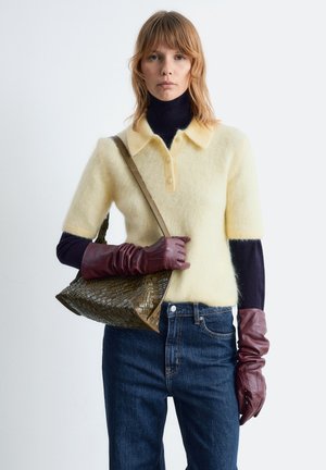 Gelb gestrickter Polo-Pullover mit kurzen Ärmeln, über einem dunklen Rollkragenpulli getragen. Kombiniert mit dunklen Jeans und bordeauxroten Lederhandschuhen. Olivgrüne Webtasche.