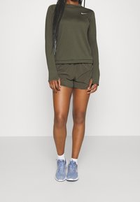 Person, der ein olivgrünes Langarmshirt und passende Shorts trägt, mit weißen Socken und hellblauen Laufschuhen vor einem schlichten Hintergrund.