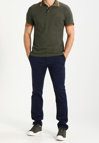 Olivgrön polo skjorta med orange detaljer och logotypapplikation, i kombination med marinblå byxor och mörka sneakers, som visar en åtsittande design.