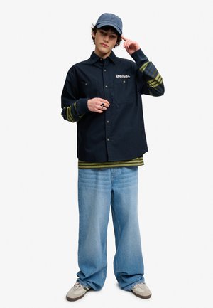 Ung person iført en blå kasket, sort knappet skjorte med "Bench" logo, stribet langærmet trøje under, brede lyseblå jeans og beige sneakers.
