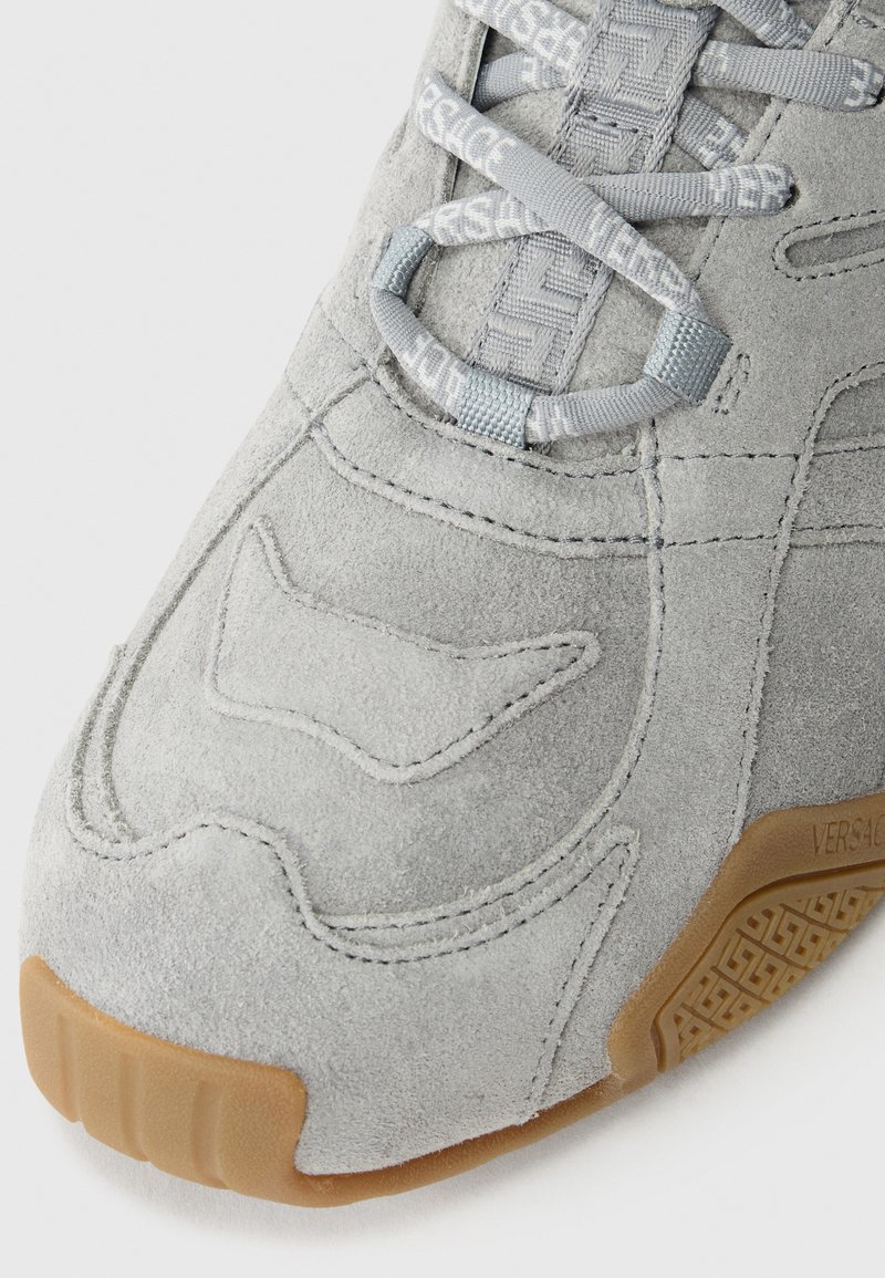 Grijze suède sneaker met een gestructureerd oppervlak, voorzien van lichtgrijze veters en een gumrubberen zool. Opvallende ontwerpelementen zijn de gestikte patronen.