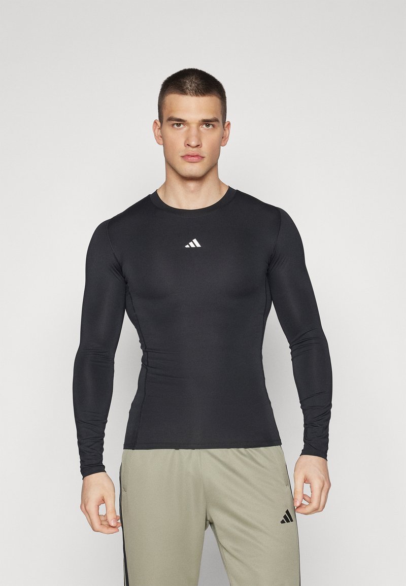 Zwart sportshirt met lange mouwen van rekbaar materiaal, met een ronde halslijn en een klein wit logo op de borst.