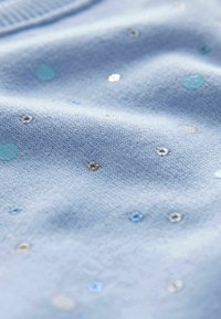 Tissu bleu clair parsemé de petits sequins brillants argentés, bleus et turquoise, montrant la texture et de doux plis.