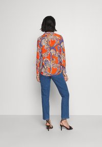 Långärmad skjorta i livlig orange med paisleymönster, kombinerad med blå raka jeans och svarta klacksandaler.