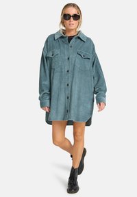 Overmaatte teal corduroy jurk met een knoopsluiting aan de voorkant, twee borstzakken en een kraag, gecombineerd met zwarte veterschoenen.