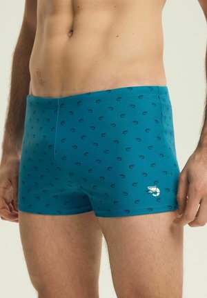 Mužský trup v tyrkysových plavkách s malými tmavými vzory krevet a bílým logem krevety poblíž levého stehna, stojící na světlém pozadí.