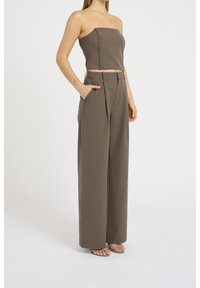 Donna che indossa un crop top brown senza spalline e pantaloni a gamba larga coordinati con dettagli a bottone lungo i lati, in piedi con una mano in tasca.