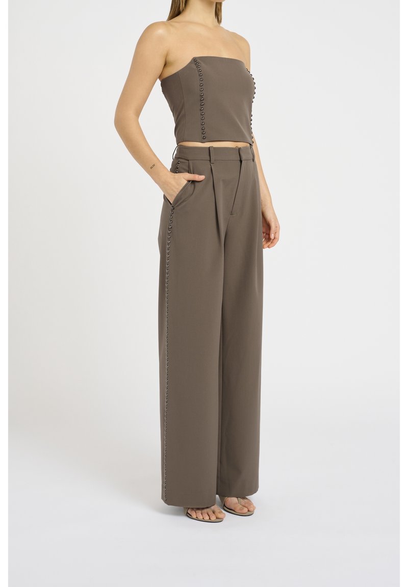 Donna che indossa un crop top brown senza spalline e pantaloni a gamba larga coordinati con dettagli a bottone lungo i lati, in piedi con una mano in tasca.