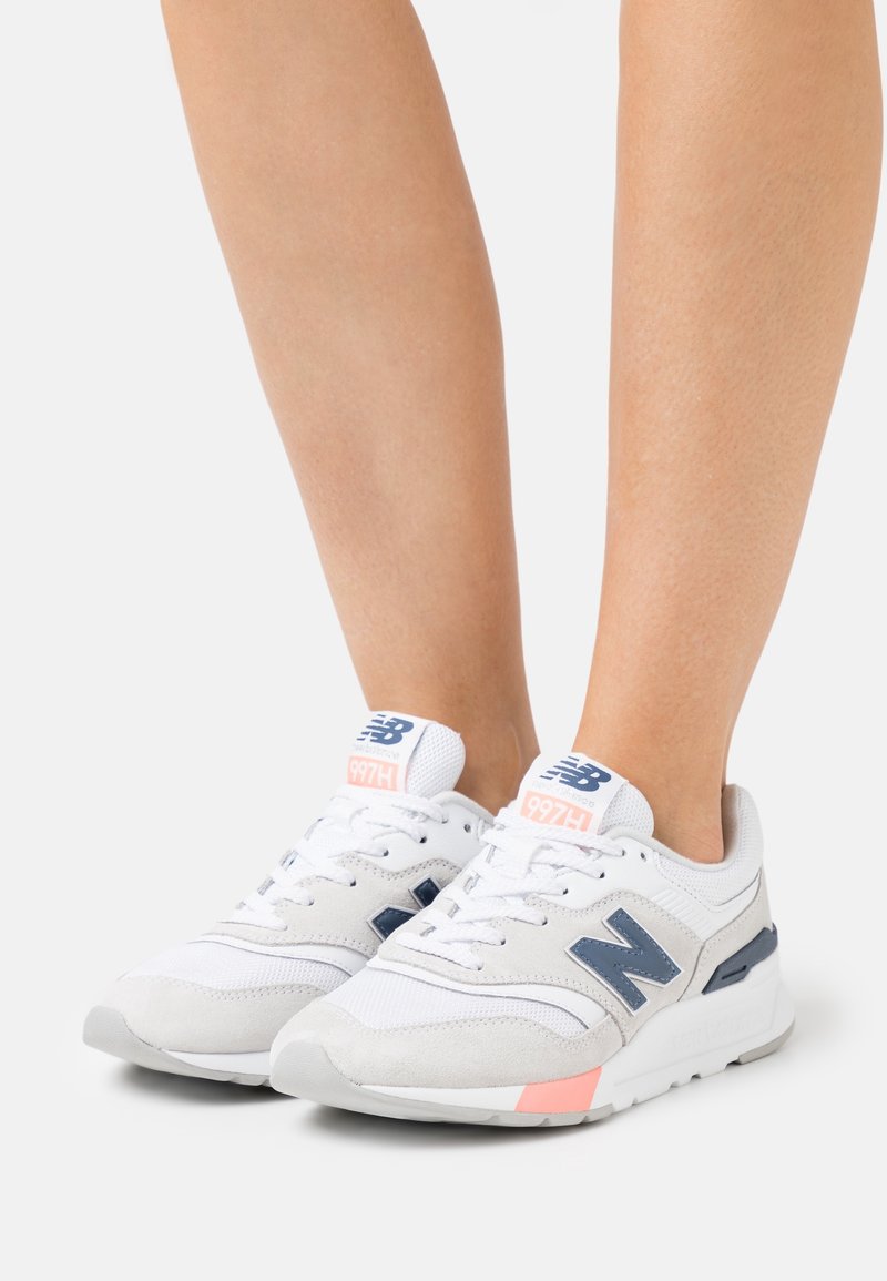Baskets blanches de sport avec des accents en daim gris et un logo bleu marine. Comprend un design à lacets, un col rembourré et un détail de talon rose.