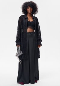 Czarna koszula na guziki z koronki nałożona na czarny, krótki top, w połączeniu z szerokimi czarnymi spodniami i torebką w kratę.