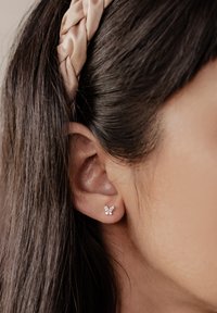 Orecchini a forma di farfalla in argento con accenti di gemme trasparenti, posti su uno sfondo di capelli lisci e scuri e una fascia di colore chiaro.