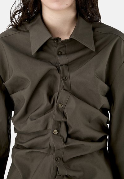 Camisa de manga larga color verde oliva oscuro con botones y tela arrugada, usada por una persona con cabello rizado y oscuro, recortada a la altura del mentón y la cintura.