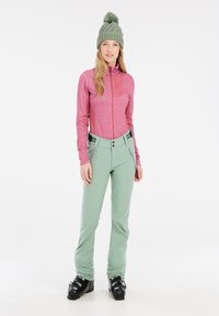 Giacca rosa aderente con zip e tasca sul petto, abbinata a pantaloni da sci verde chiaro con ferramenta nera e vita alta. Stivali da sci neri.