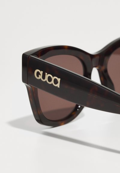 Tortoiseshell zonnebril met bruine lenzen, voorzien van een gedurfde frameontwerp en gouden "GUCCI" logo op de poot. Glad textuur en hoekige vorm.