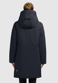 Tumedat sinine, põlvini ulatuv naiste puffer-kurta suure kapuutsiga, küljeosal tõmblukuga taskuga ja tekstureeritud kangaga. Logodetail seljal.