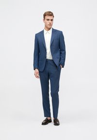 Costum slim-fit de culoare albastră, confecționat dintr-un material texturat, având un sacou cu un singur rând de nasturi și pantaloni asortați, purtat cu o cămașă albă și pantofi maro.