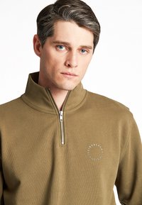 Felpa verde oliva con mezza zip e finitura texturizzata, caratterizzata da un piccolo logo circolare sul lato sinistro del petto e un colletto strutturato.