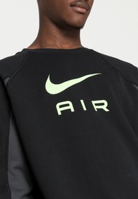 Svart sweatshirt med rund halsringning, prydd med en ljusgrön Nike Swoosh-logotyp och texten "AIR" på framsidan. Har grå accenter på ärmarna.