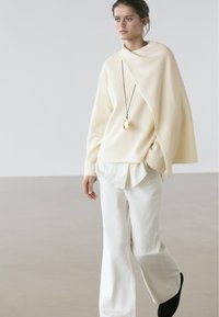 Cream-colored, oversized sweater with draped design, layered over a white shirt, paired with wide-leg white pants and black shoes. Gold pendant accessory.
