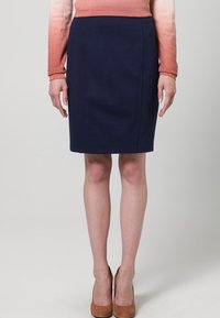 Jupe crayon bleu marine foncé au toucher lisse et à la coupe structurée, agrémentée de détails de coutures subtils. Portée avec des talons de couleur claire.