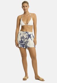 Een vrouw staat zelfverzekerd, gekleed in een witte bikini-top en bloemen korte broek, met een neutrale achtergrond die haar ontspannen zomerse stijl benadrukt.
