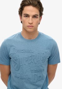 Camiseta de algodón azul claro con el logotipo texturizado de "Superdry" en un diseño en relieve, mangas cortas y cuello redondo.