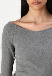 Donna che indossa un maglione grigio a maniche lunghe aderente e a costine con un ampio scollo off-the-shoulder che mette in mostra le clavicole.