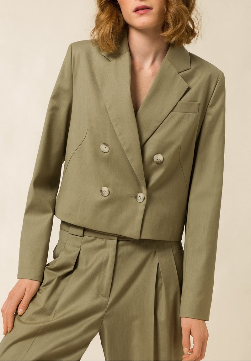 Femme portant un blazer court olive à double boutonnage et un pantalon plissé taille haute assorti sur fond beige.
