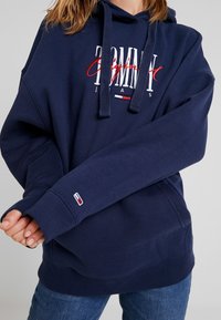 Mörkblå hoodie med stort vitt och rött "Tommy Jeans"-logotyp. Har dragsko, ribbade muddar och en liten logotyp på ärmen.