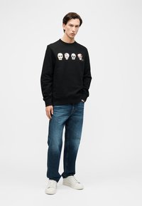 Νεαρός άνδρας που στέκεται φορώντας μαύρο φούτερ με τέσσερα πολύχρωμα σχέδια κρανίων, μπλε τζιν και λευκά αθλητικά παπούτσια, μπροστά σε απλό φόντο.