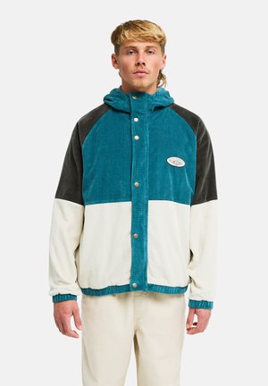 Giovane uomo con capelli biondi che indossa una giacca di velluto a coste a blocchi di colore blu, nero e bianco con bottoni a pressione e pantaloni beige.