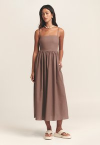 Maxiklänning i enfärgat brunt, tillverkad av mjukt tyg. Har en fyrkantig halsringning, tunna band och en rynkad midja med sidosfickor.