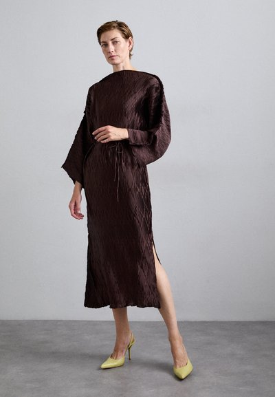 Woman with short hair wearing a textured dark brown midi dress with wide sleeves and yellow high-heeled shoes, standing against a plain gray wall.